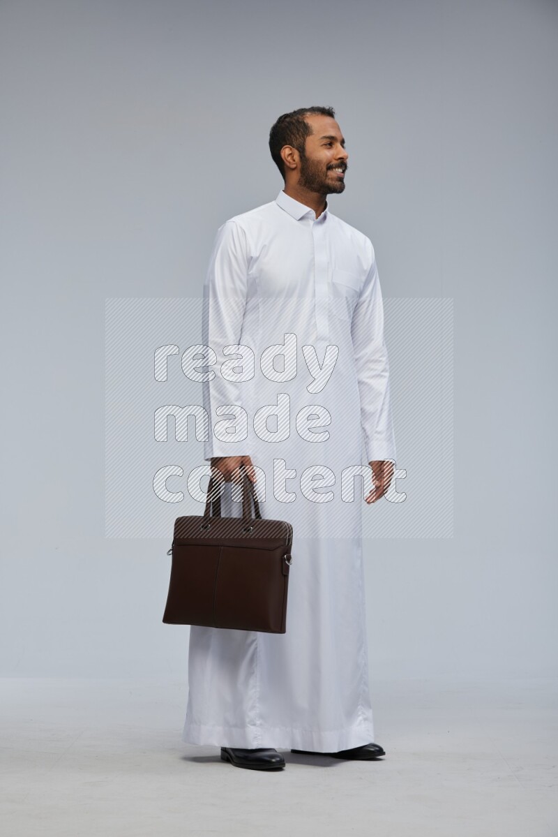Saudi man wearing thob standing holding suitcase on gray background