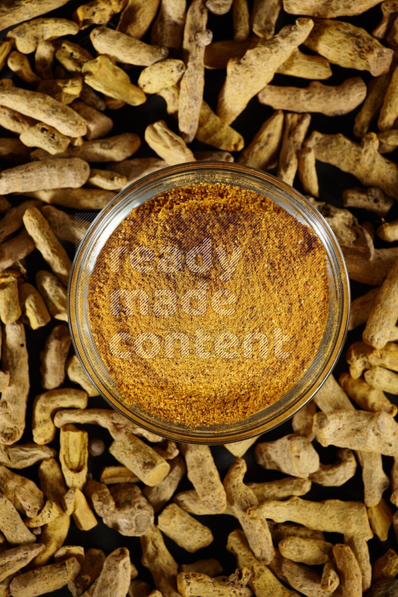 A glass bowl full of turmeric powder and dried whole fingers beside it on black flooring