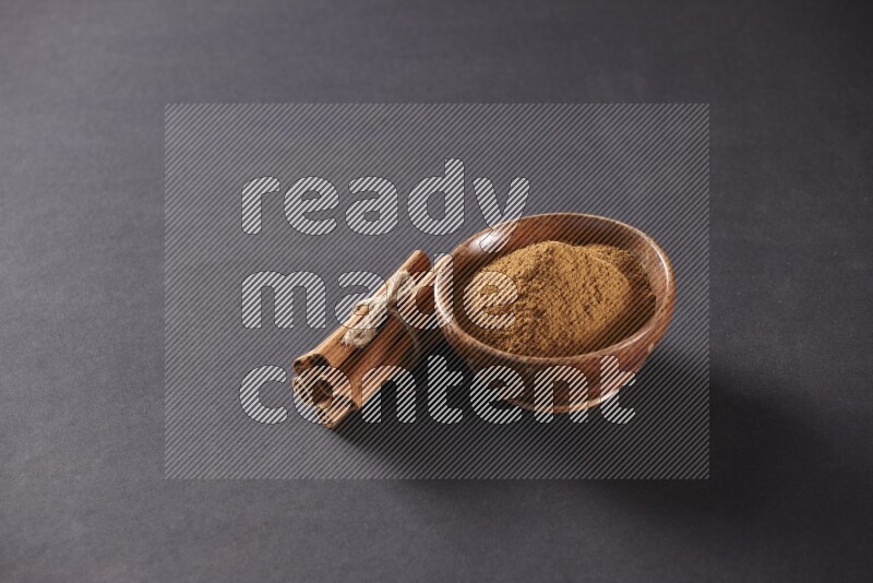 Cinnamon sticks stacked and bounded beside a wooden bowl full of cinnamon powder and a wooden spoon full of powder on black background