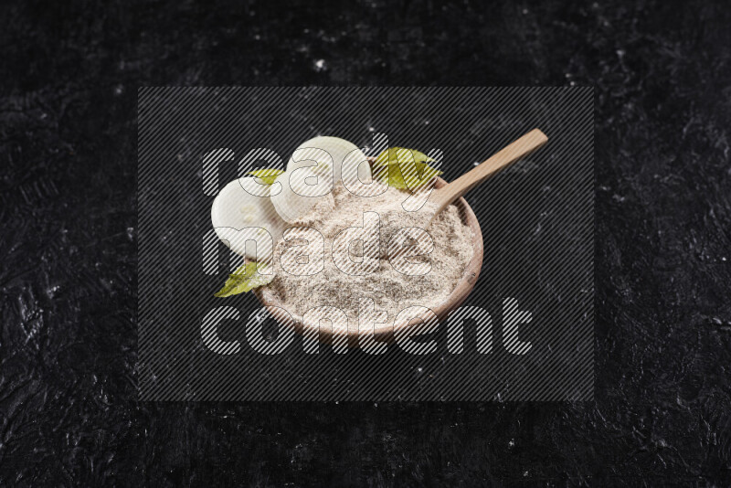 A wooden bowl full of onion powder with a wooden spoon in it with some sliced onions on black background