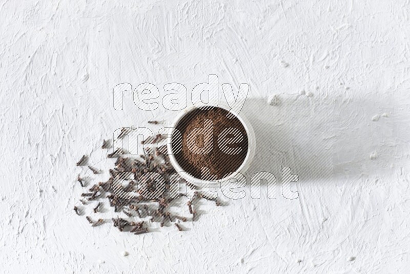 A beige ceramic bowl full of cloves powder and whole cloves on a white flooring