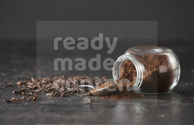 A flipped glass spice jar and a metal spoon full of cloves powder with cloves spread on textured black flooring
