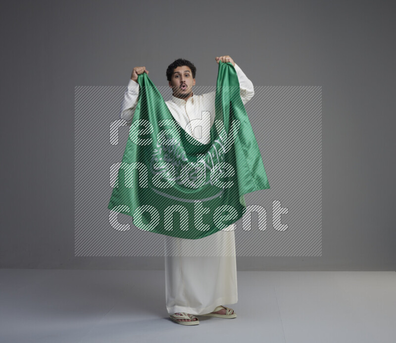 A Saudi man standing wearing thob holding big Saudi flag on gray background