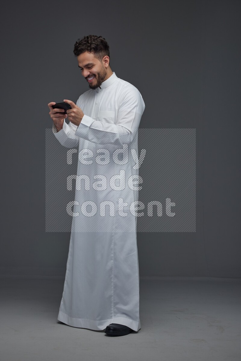 A Saudi man wearing Thobe and playing using his phone horizontally playing video games while standing and making different poses eye level on a gray background