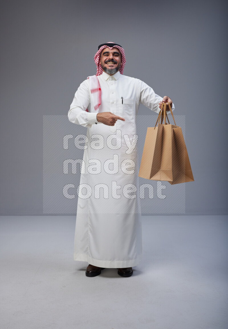 Saudi man Wearing Thob and red Shomag standing holding shopping bag on Gray background