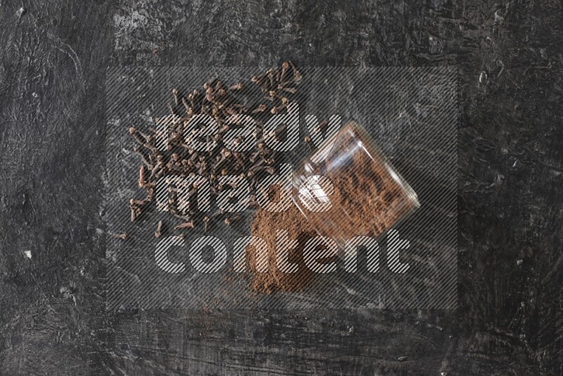 وعاء زجاجي مقلوب ممتلئ ببودرة القرنفل مع حبوب القرنفل المتناثرة على أرضية سوداء