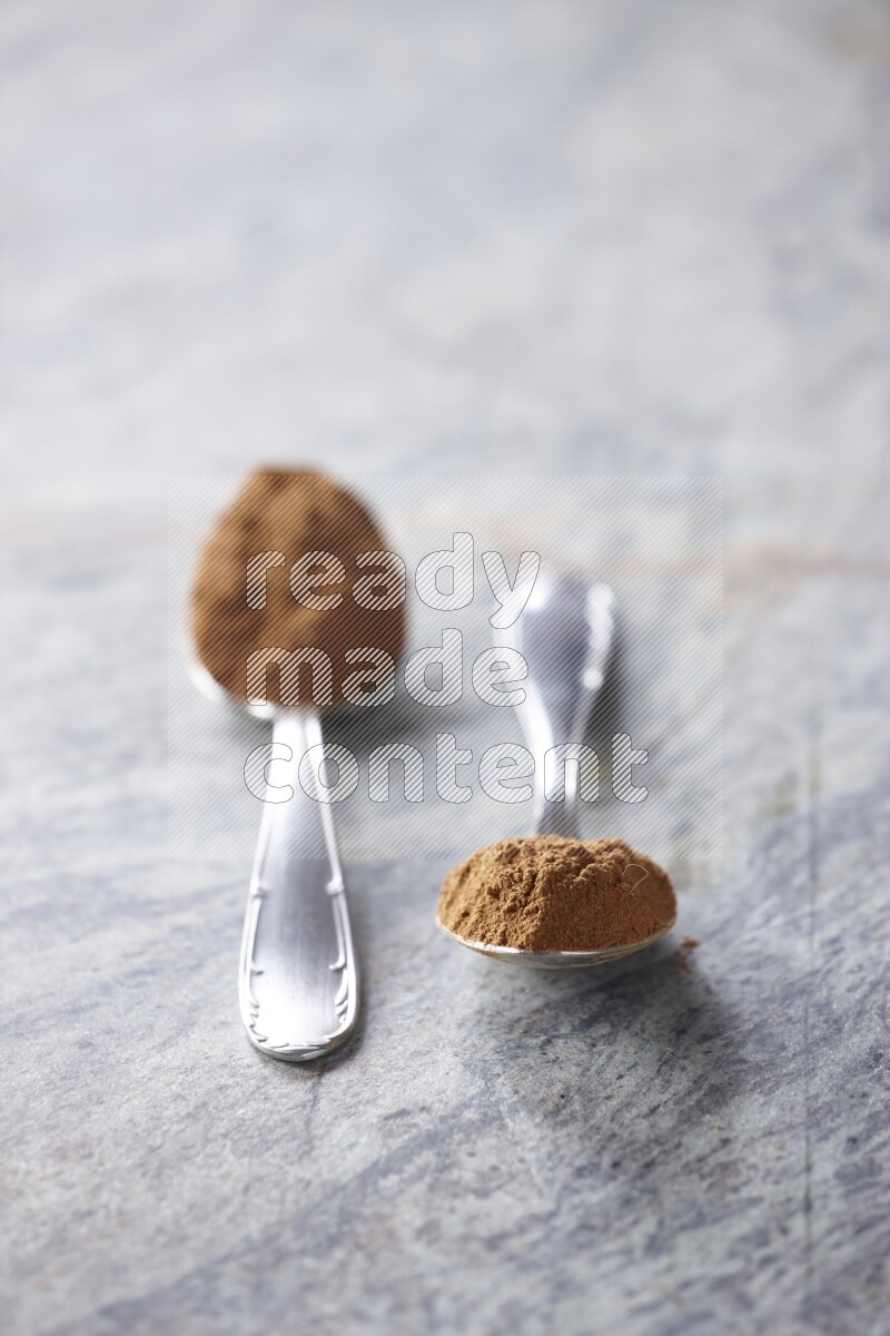 Two metal spoons full of cinnamon powder on grey marble background