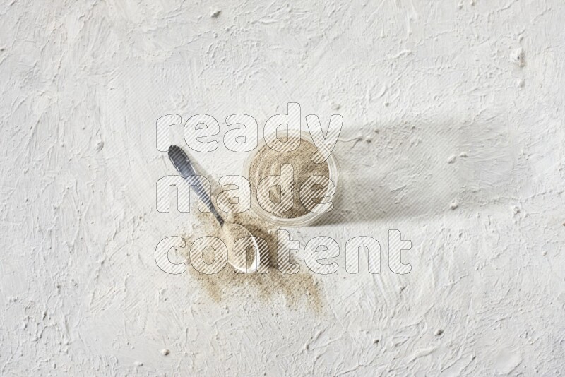 A glass jar and a metal spoon full of white pepper powder on textured white flooring