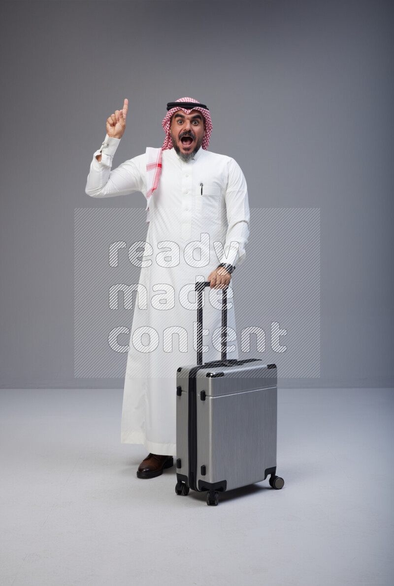 Saudi man wearing Thob and red Shomag standing holding Travel bag on Gray background
