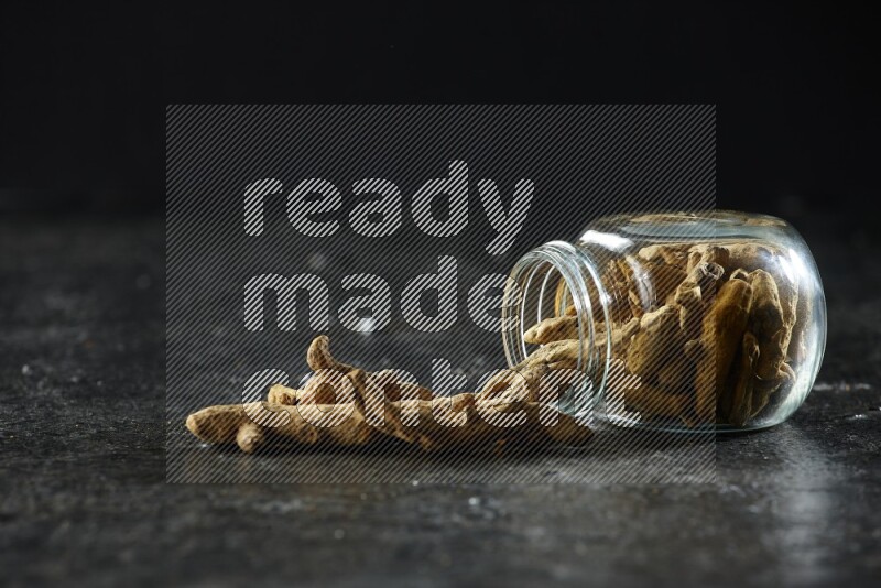 A flipped glass spice jar full of dried turmeric whole fingers and fingers fell out of it on textured black flooring