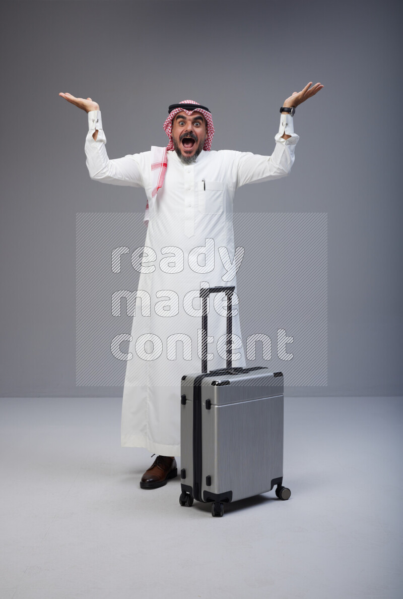Saudi man wearing Thob and red Shomag standing holding Travel bag on Gray background