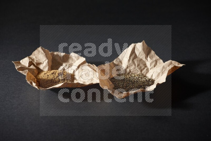 Cumin seeds and powder in 2 crumpled pieces of paper on black flooring