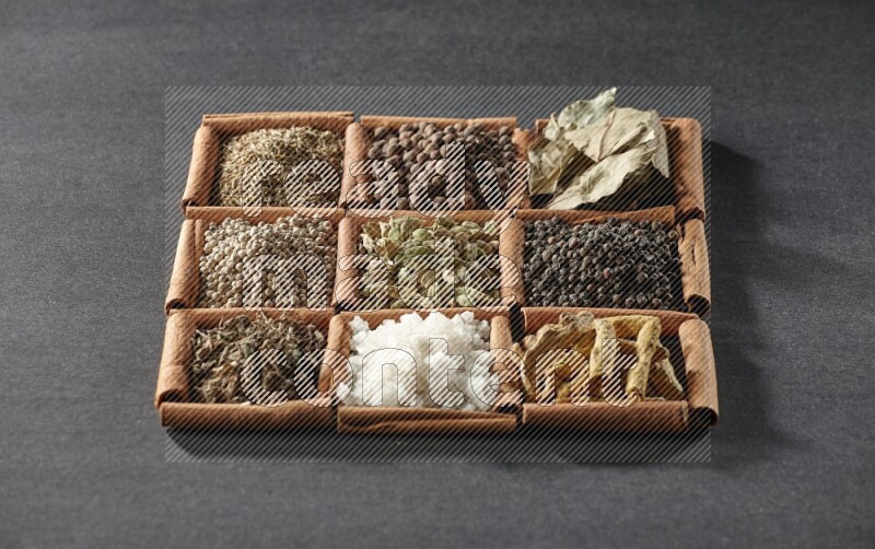 9 squares of cinnamon sticks full of cardamom in the middle surrounded by turmeric, black peppers, bay laurel, allspice, cumin, white peppers, basil and salt on black flooring