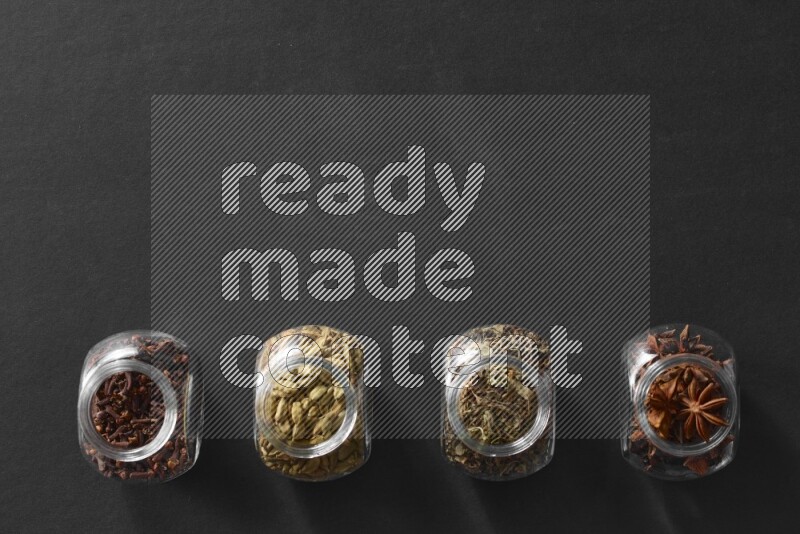4 glass spice jars full of cloves, cardamom, basil and star anise on black flooring