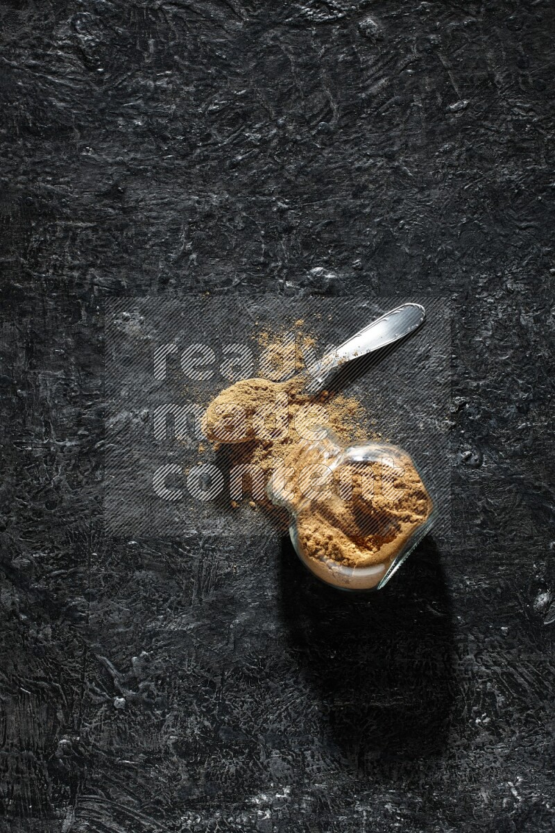 وعاء زجاجي للتوابل مقلوب وملعقة معدنية ممتلئان ببودرة بهار الفلفل الحلو والبودرة تناثرت من الوعاء على خلفية سوداء