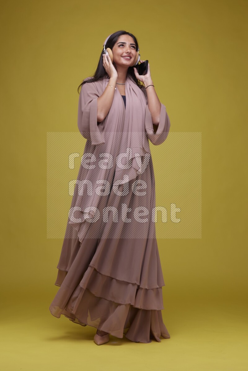 A Saudi woman Listing to Music on a Yellow Background wearing Brown Abaya