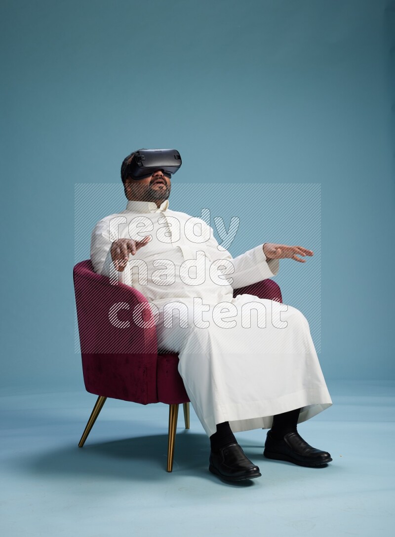Saudi Man without shimag sitting on chair  gaming with VR on blue background