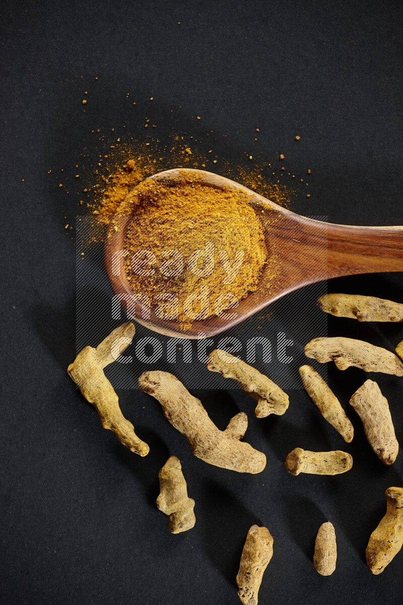 A wooden ladle full of turmeric powder with dried turmeric fingers on black flooring