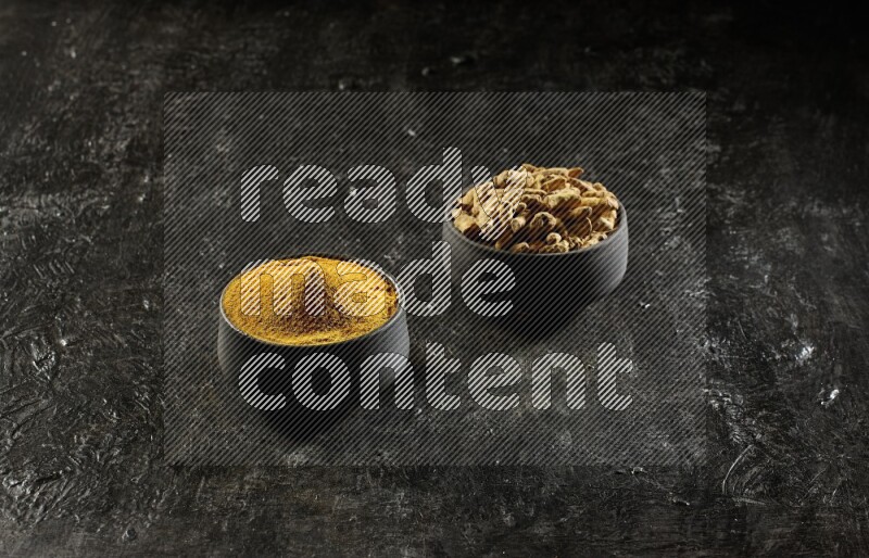 2 pottery black bowls, one full of turmeric powder and the other full of dried turmeric whole fingers on a textured black flooring