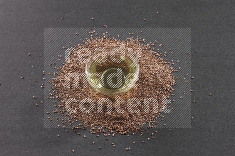 A glass bowl full of flaxseeds oil surrounded by the seeds on a black flooring