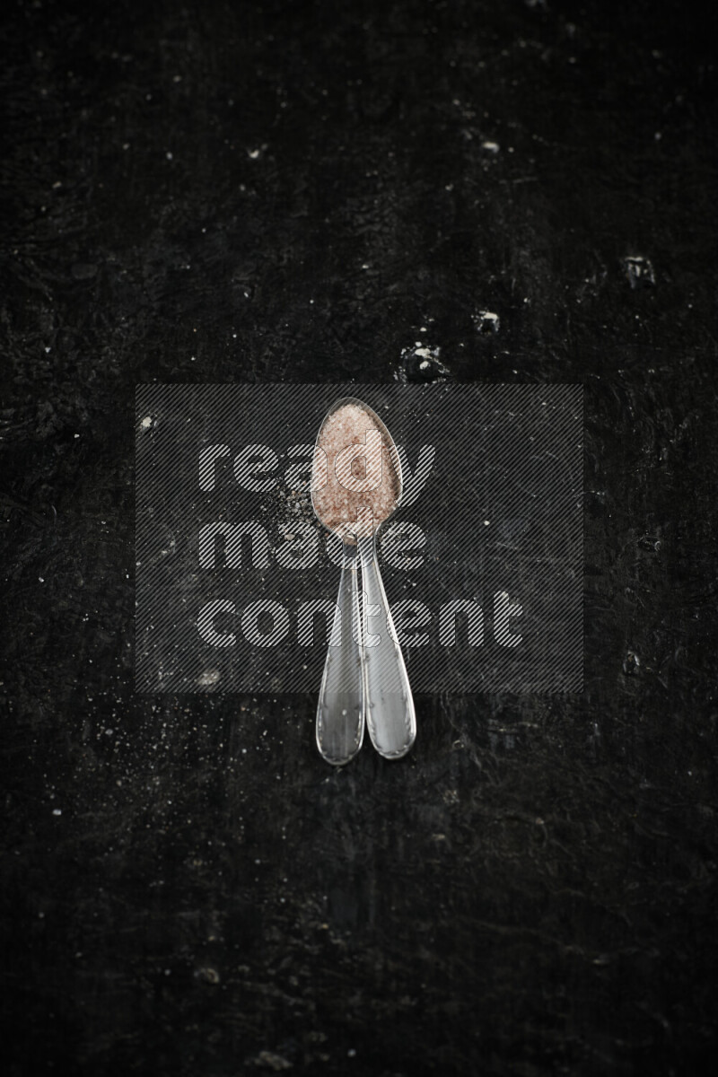 2 metal spoons filled with pink himalayan salt on black background