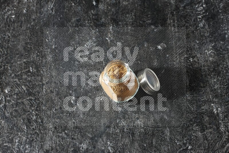A glass spice jar full of allspice powder on a textured black flooring