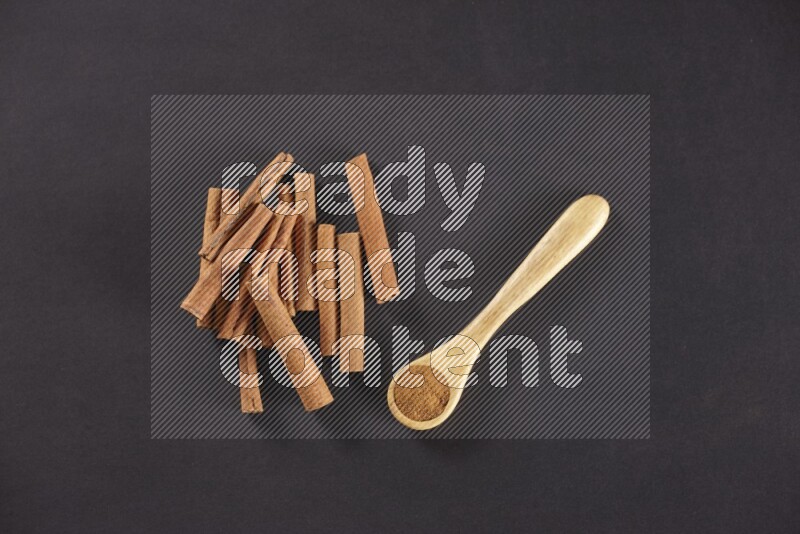 Cinnamon sticks stacked beside a wooden spoon full of cinnamon powder on black background