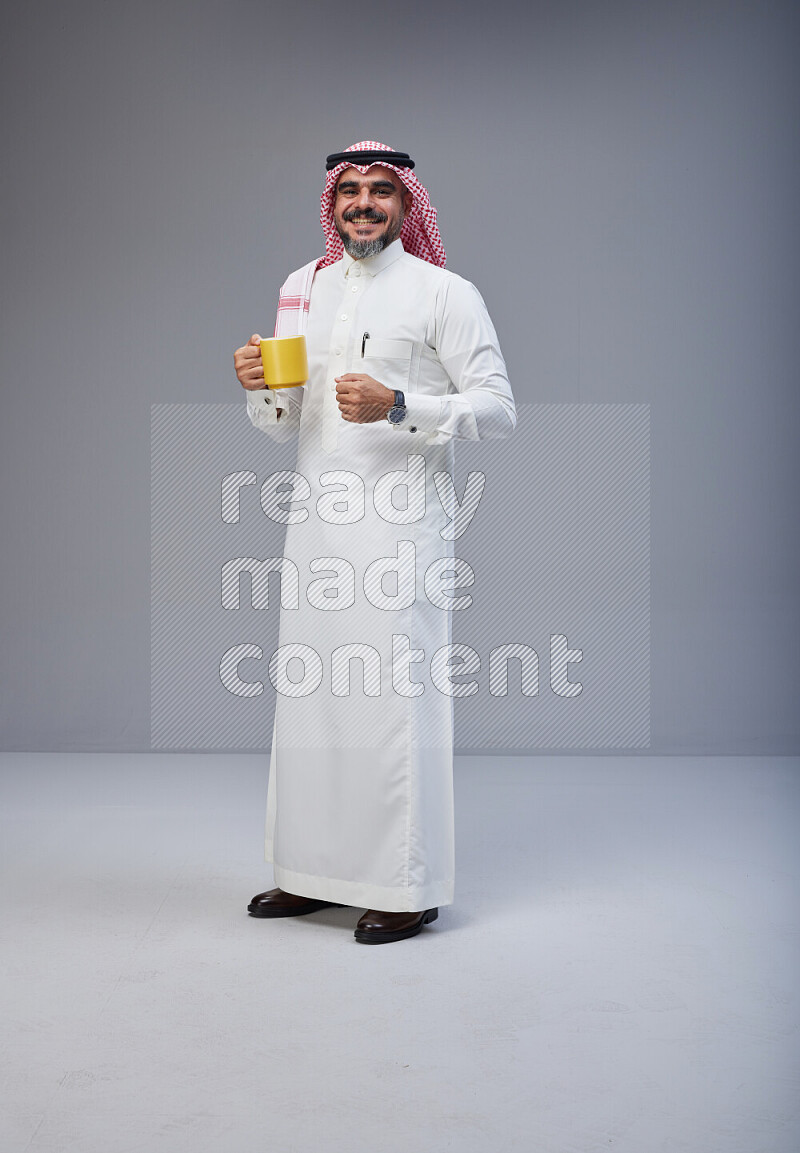 Saudi man Wearing Thob and red Shomag standing holding mug on Gray background