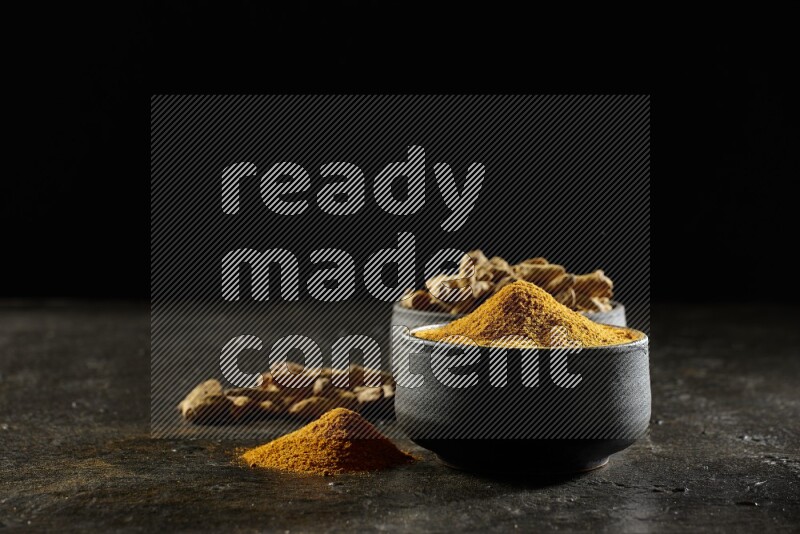 2 pottery black bowls, one full of turmeric powder and the other full of dried turmeric whole fingers on a textured black flooring