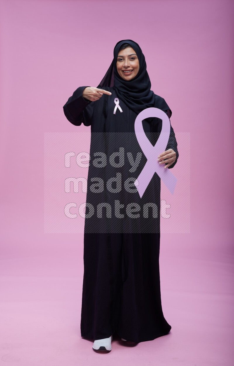 Saudi woman wearing pink ribbon on abaya standing holding awareness ribbon on plain pink background