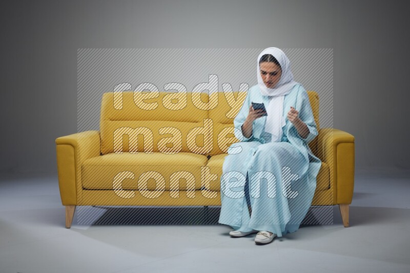 A Saudi woman wearing a light blue Abaya and white head scarf sitting on a yellow sofa and texting on her phone on a grey background