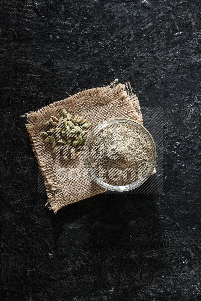 A glass bowl full of cardamom powder with cardamom seeds on a burlap piece on textured black flooring