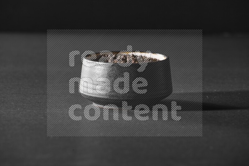 A black pottery bowl full of black pepper beads on black flooring