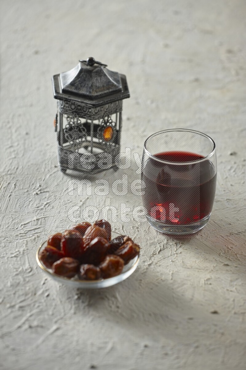 A silver lantern with different drinks, dates, nuts, prayer beads and quran on textured white background