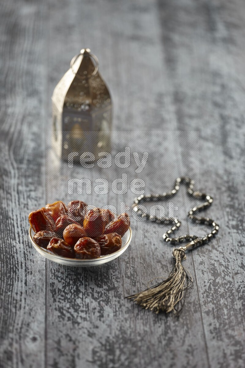A silver lantern with different drinks, dates, nuts, prayer beads and quran on grey wooden background