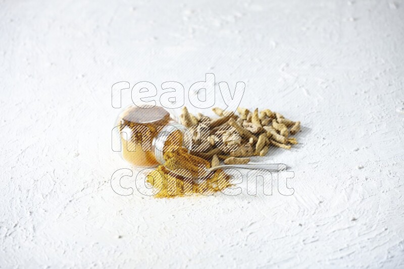 وعاء زجاجي للتوابل مقلوب وملعقة معدنية ممتلئة ببودرة الكركم والبودرة تناثرت منه مع أصابع الكركم المجففة على خلفية بيضاء