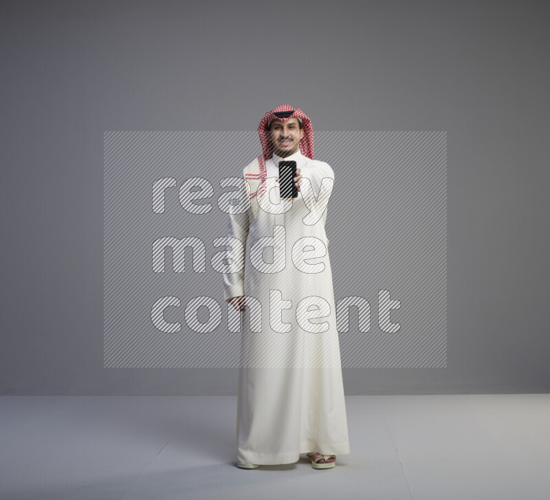 A Saudi man standing wearing thob and red shomag showing phone to camera on gray background