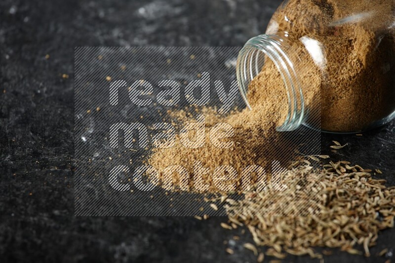 A flipped glass spice jar full of cumin powder with spilled powder and cumin seeds on a textured black flooring