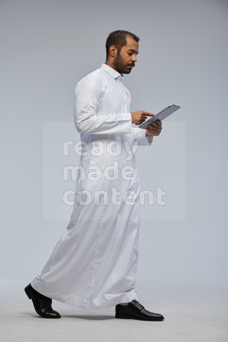 Saudi man wearing Thob standing working on tablet on Gray background