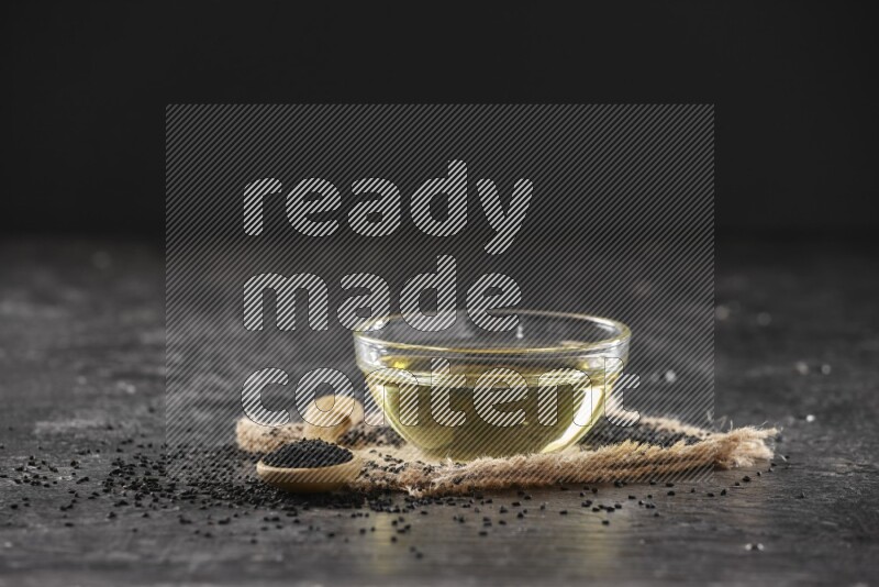 A glass bowl full of black seeds oil and wooden spoon full of black seeds with seeds spreaded on burlap fabric on a textured black flooring