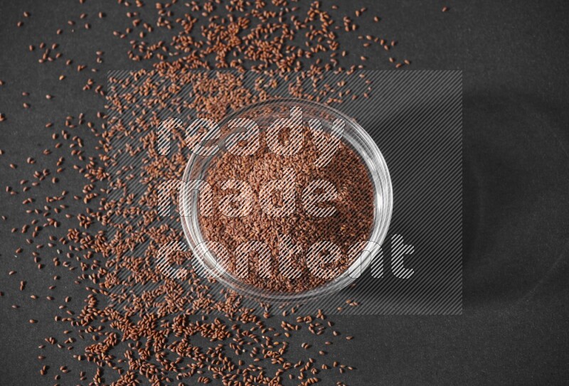A glass bowl full of garden cress seeds surrounded by seeds on a black flooring