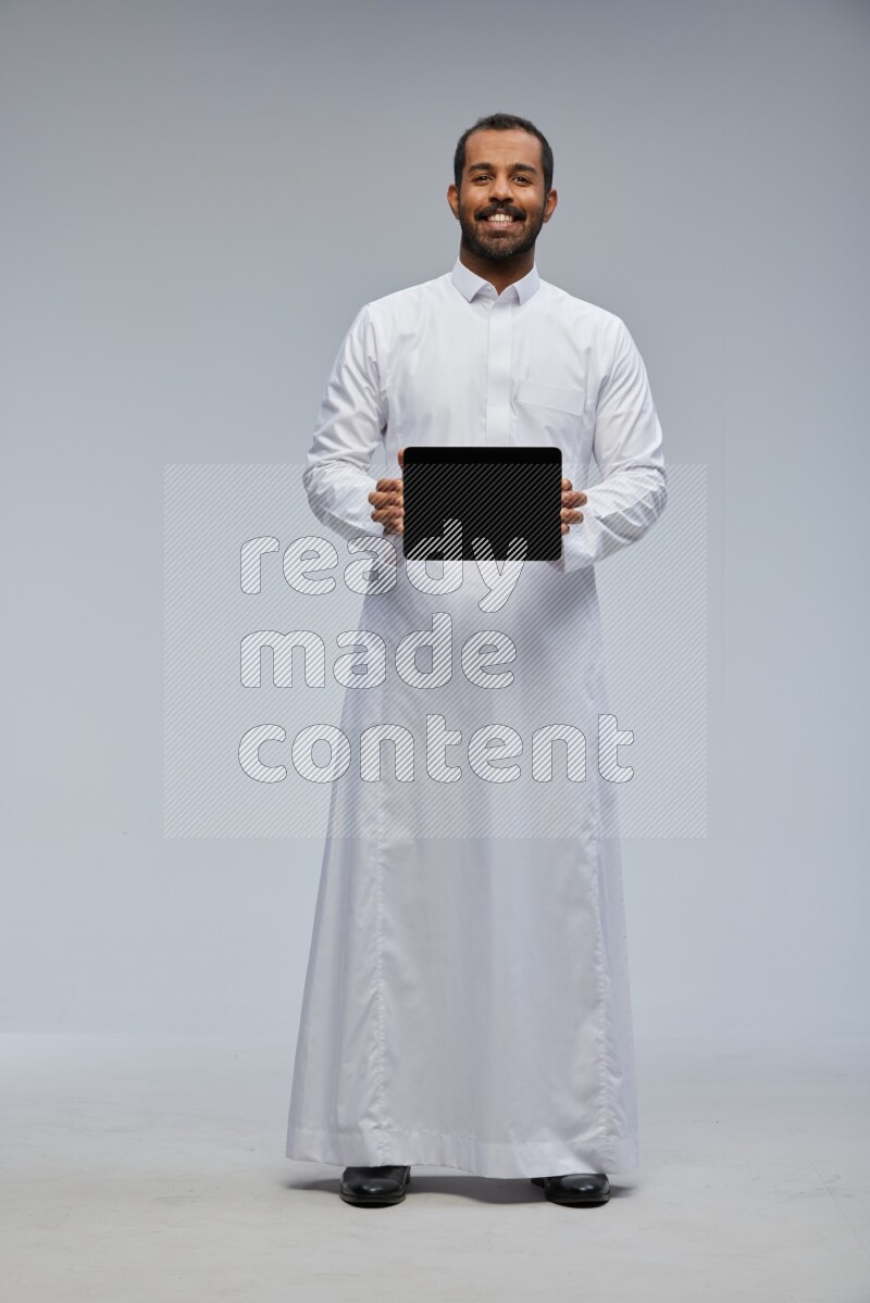 Saudi man wearing Thob standing showing tablet to camera on Gray background