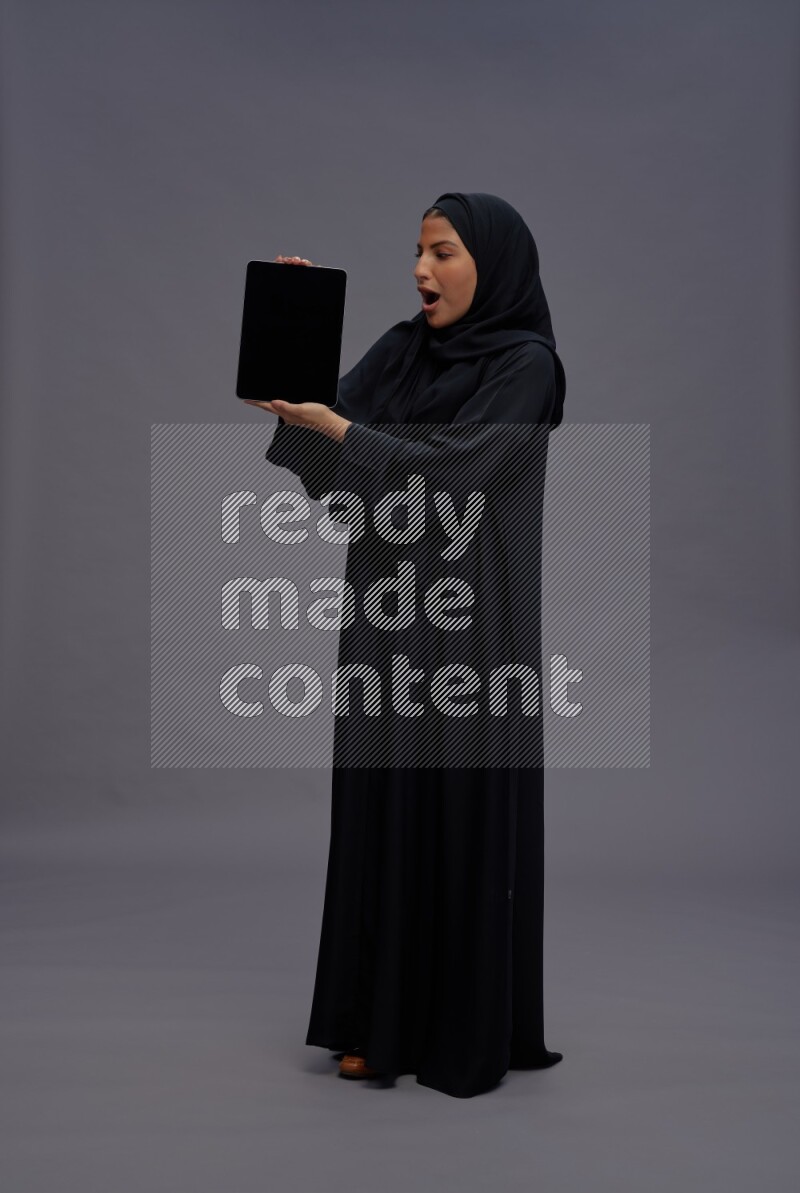 Saudi woman wearing Abaya standing showing tablet to camera on gray background