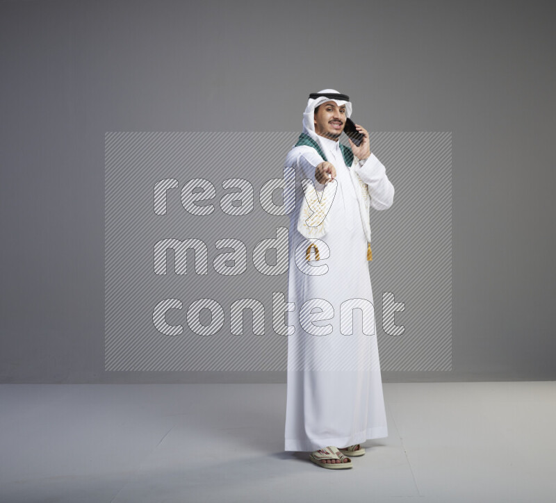 A saudi man standing wearing thob and white shomag with flag scarf on his neck talking on phone on gray background