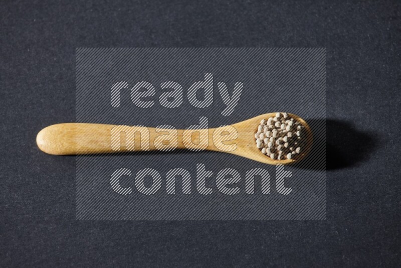 A wooden spoon full of white pepper beads on black flooring