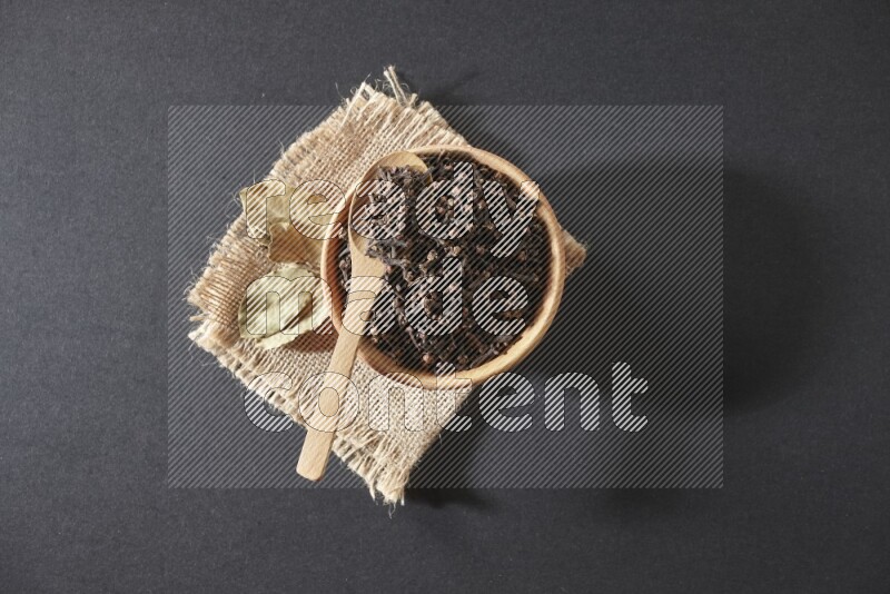 A wooden bowl, a wooden spoon full of cloves, and bay leaves (laurel) on a piece of burlap on a black flooring