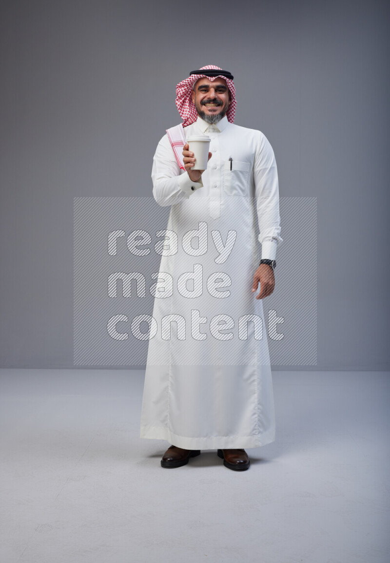 Saudi man Wearing Thob and red Shomag standing holding paper cup on Gray background