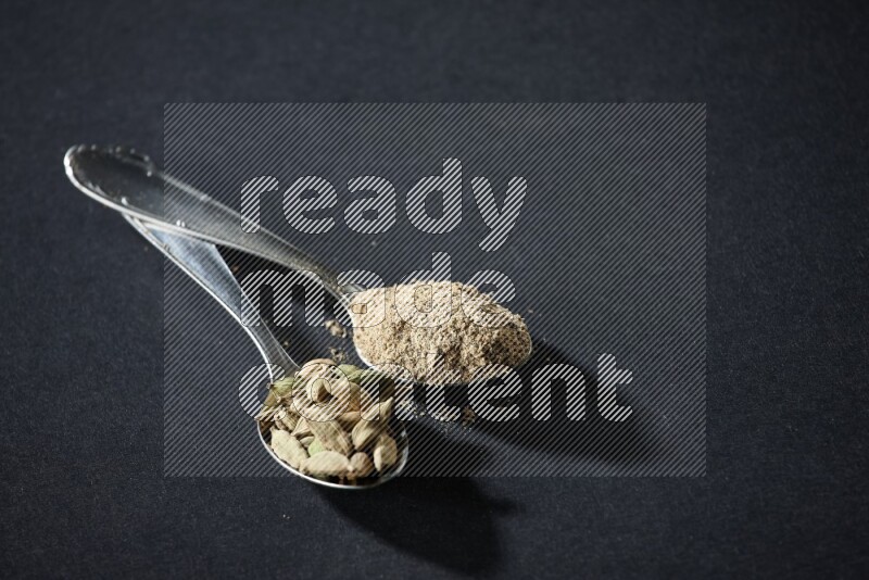 ملعقتان معدنيتان ممتلئتان ببودرة الهيل وحبوب الهيل على خلفية سوداء