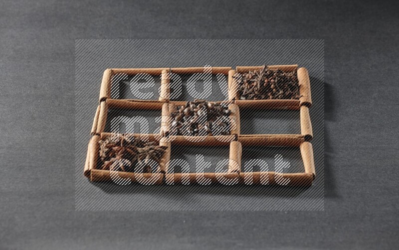 9 squares of cinnamon sticks full of coffee beans in the middle surrounded by nutmeg, dried mint, cloves, dried basil, dried ginger, cinnamon, star anise and cardamom on black flooring
