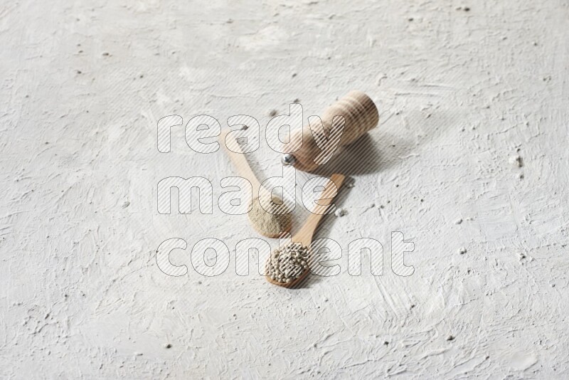 2 wooden spoons one full of white pepper powder and the other with pepper beads and a wooden pepper grinder on textured white flooring